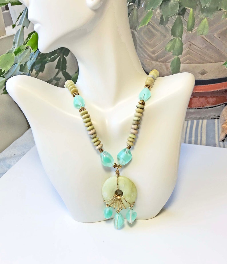 May include: A handcrafted necklace featuring a large, pale green circular pendant with a gold wire design and dangling aqua-colored beads. The necklace has light green and tan wooden beads and aqua-colored glass beads.