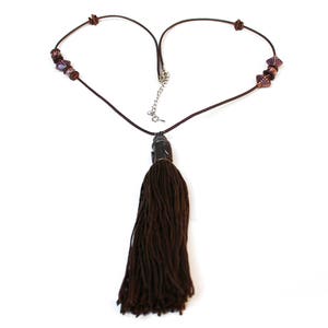 May include: A brown necklace with a long tassel pendant. The necklace cord forms a heart shape. The necklace has small, dark purple beads on each side of the heart shape. The tassel is made of thick, dark brown threads.