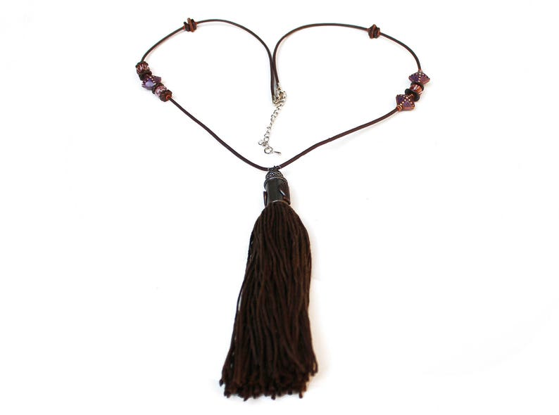 May include: A brown necklace with a long tassel pendant. The necklace cord forms a heart shape. The necklace has small, dark purple beads on each side of the heart shape. The tassel is made of thick, dark brown threads.