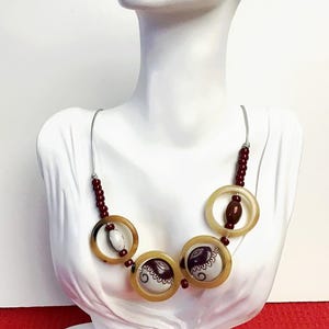 May include: A necklace featuring three large translucent tan circular rings, two of which contain white and dark brown patterned circular inserts. The piece includes small dark red beads and a white oval bead, all strung on a thin silver-toned wire.