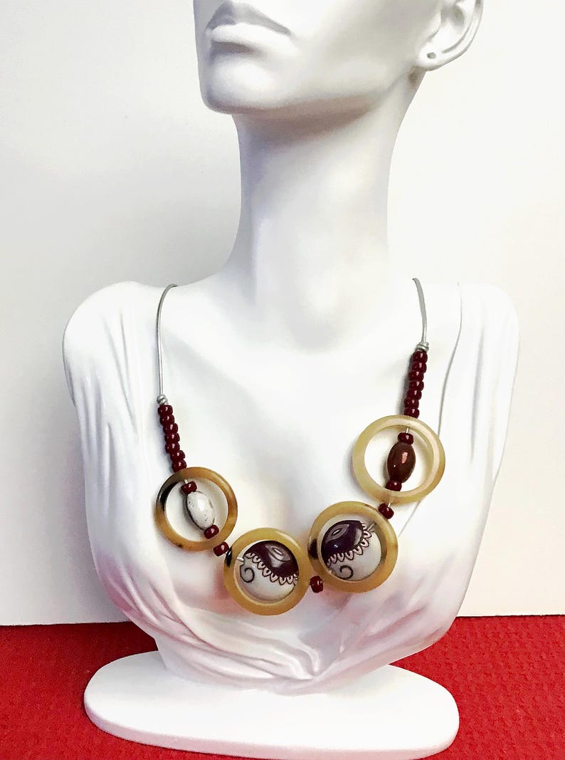 May include: A necklace featuring three large translucent tan circular rings, two of which contain white and dark brown patterned circular inserts. The piece includes small dark red beads and a white oval bead, all strung on a thin silver-toned wire.