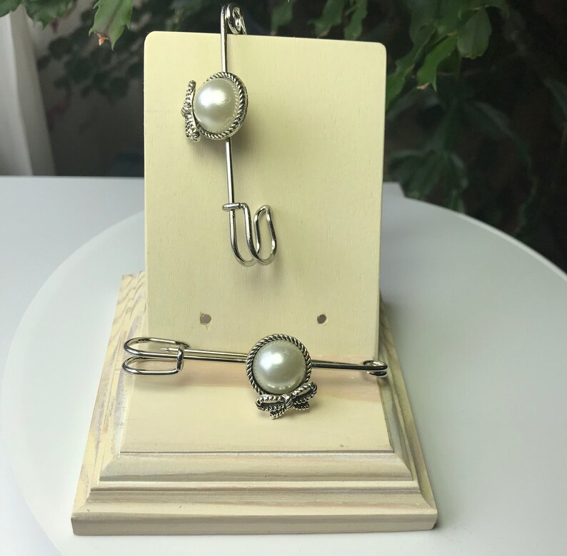 May include: Two silver-toned page holders with a pearl-like bead and decorative bow. The page holders are displayed on a cream-colored wooden stand with a decorative base. The design is elegant and suitable for books or sheet music.