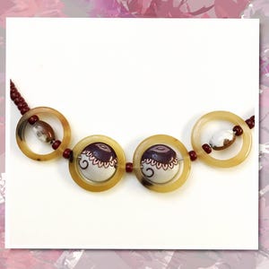 May include: A necklace featuring four large, translucent, amber-colored circular rings. The two central rings contain white and dark purple patterned ceramic beads, while the outer rings hold smaller, mottled brown and white oval beads.