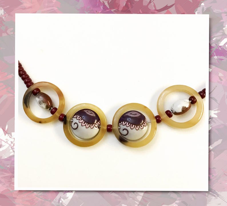 May include: A necklace featuring four large, translucent, amber-colored circular rings. The two central rings contain white and dark purple patterned ceramic beads, while the outer rings hold smaller, mottled brown and white oval beads.