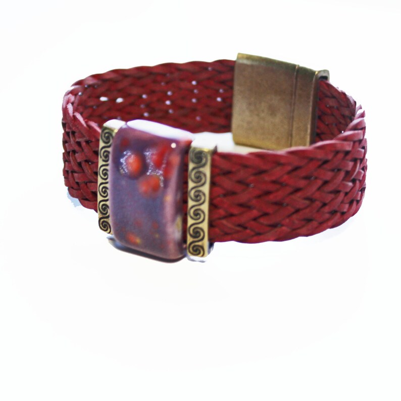 May include: A braided red leather bracelet with a rectangular, speckled, purple and red centerpiece. The centerpiece is framed with gold-colored metal and has a magnetic clasp. The bracelet is a stylish accessory.
