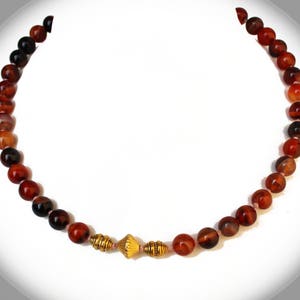 May include: A beaded necklace featuring reddish-brown and black round beads. The necklace has a gold-colored, decorative centerpiece. The beads are strung together, creating a circular shape. The necklace is displayed against a white background.