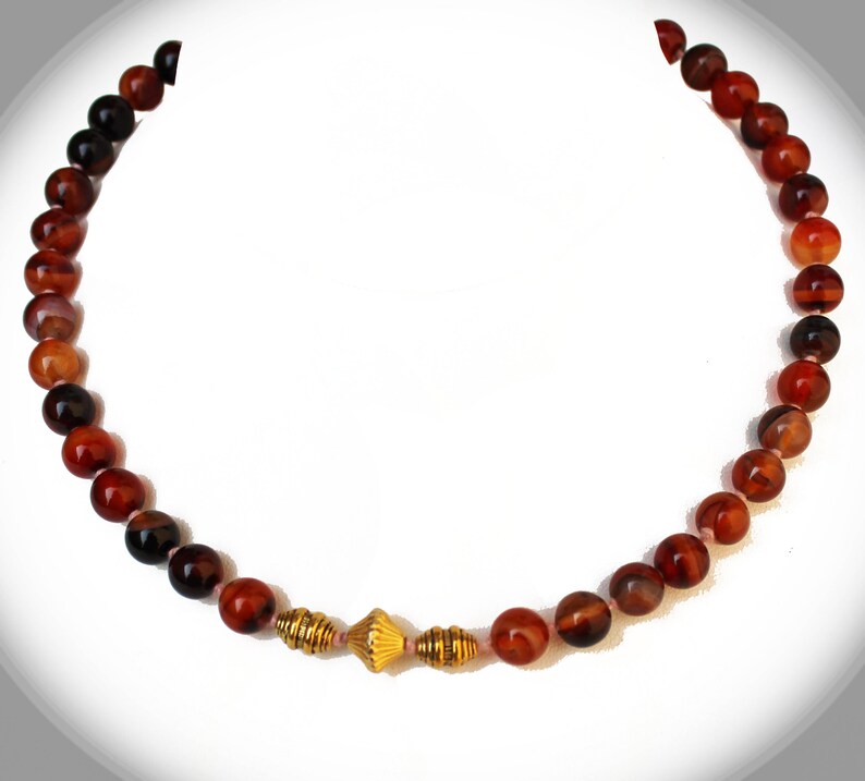 May include: A beaded necklace featuring reddish-brown and black round beads. The necklace has a gold-colored, decorative centerpiece. The beads are strung together, creating a circular shape. The necklace is displayed against a white background.