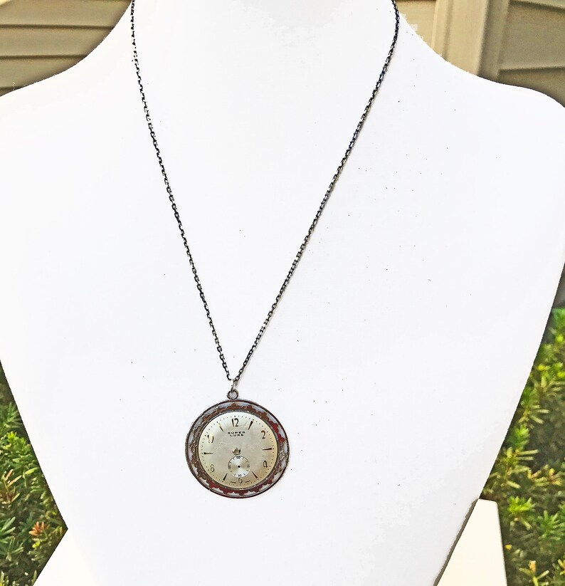 May include: A vintage watch face pendant necklace. The round watch face has a silver-tone dial with black numerals and hands. The bezel is a mix of red and silver. The pendant hangs from a dark chain, displayed on a white mannequin.