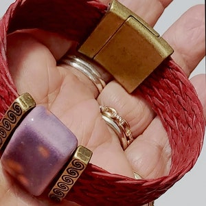 May include: Red braided leather bracelet with a square purple bead and gold-colored metal accents. The bracelet features a magnetic clasp and decorative metal details. The bracelet is approximately 7 inches in length.