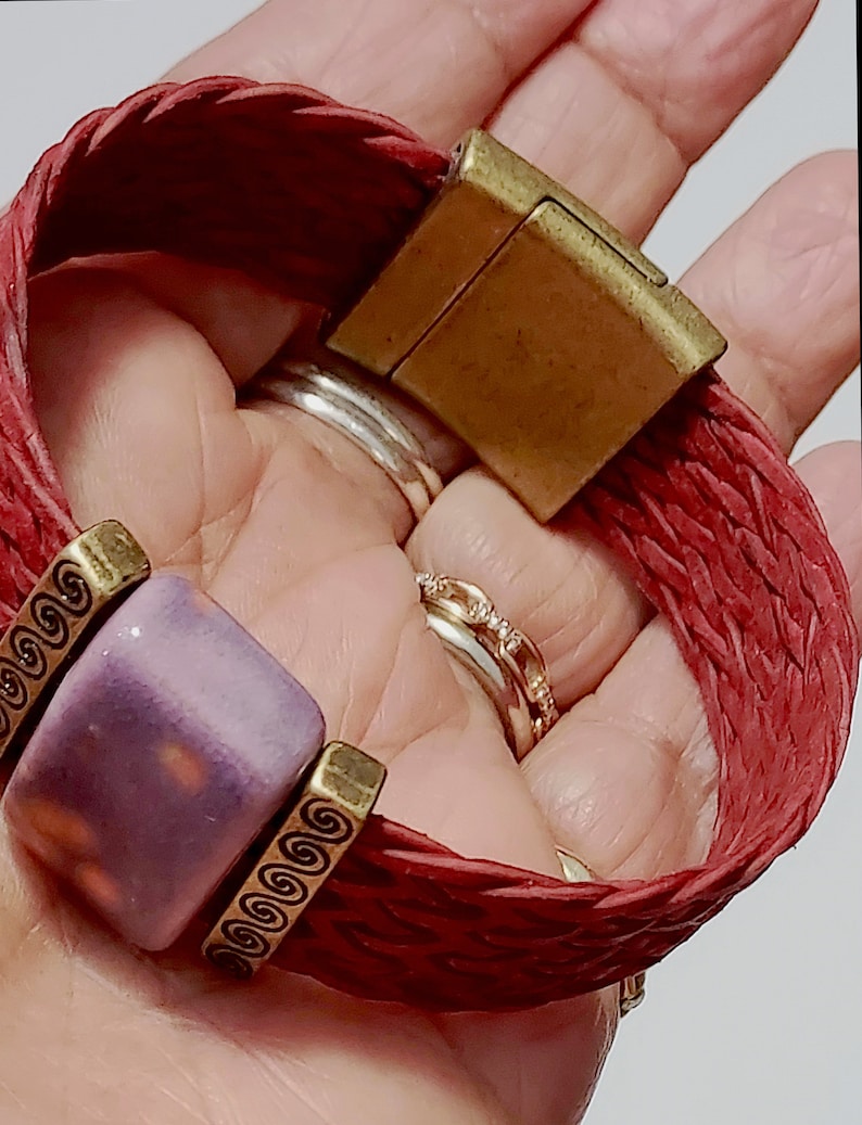 May include: Red braided leather bracelet with a square purple bead and gold-colored metal accents. The bracelet features a magnetic clasp and decorative metal details. The bracelet is approximately 7 inches in length.