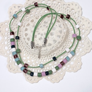 May include: A green leather necklace with colorful square beads. The beads are in shades of white, green, purple, blue, and dark red. The necklace is displayed on a white lace doily.