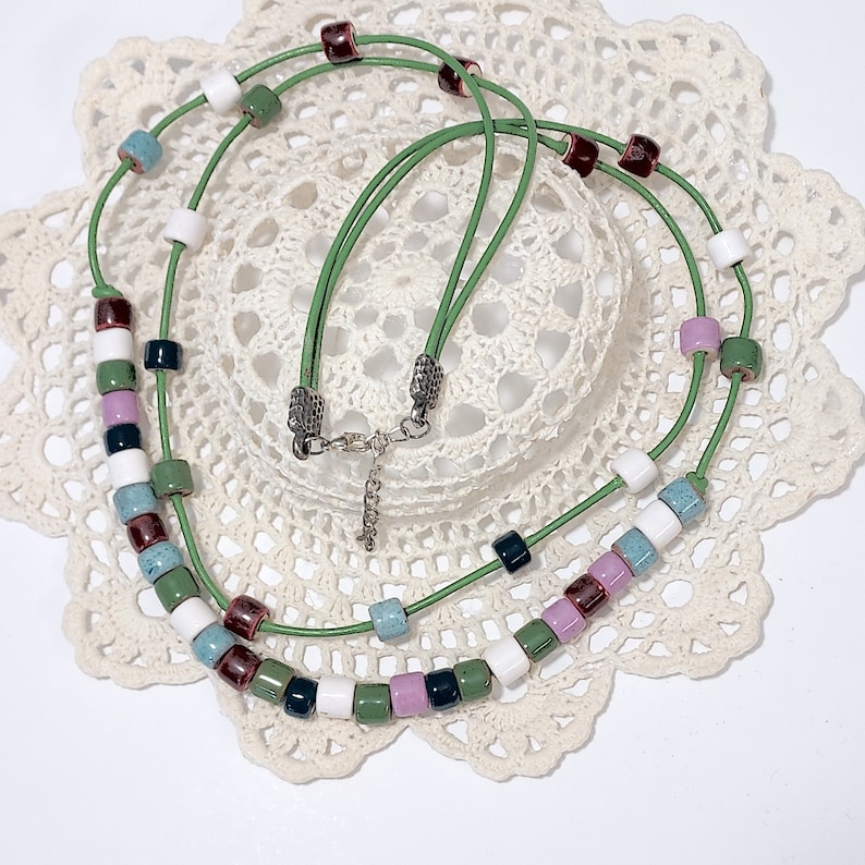 May include: A green leather necklace with colorful square beads. The beads are in shades of white, green, purple, blue, and dark red. The necklace is displayed on a white lace doily.
