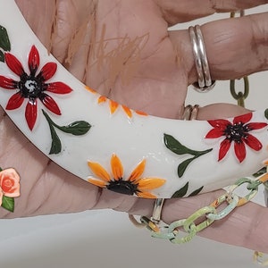 May include: A white crescent-shaped necklace pendant adorned with red and orange flower designs and green leaves. The pendant is connected to a colorful chain. The necklace is held by a person wearing silver rings.