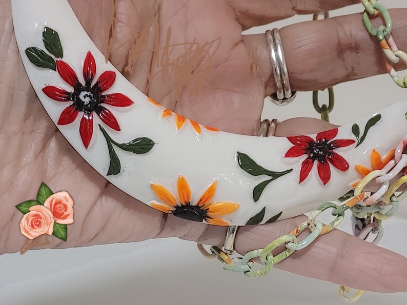 May include: A white crescent-shaped necklace pendant adorned with red and orange flower designs and green leaves. The pendant is connected to a colorful chain. The necklace is held by a person wearing silver rings.