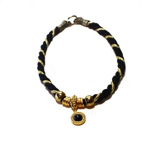 May include: A black and gold rope necklace with a gold pendant featuring a black stone. The necklace has a twisted rope design with gold accents. The pendant is round and has a gold frame.