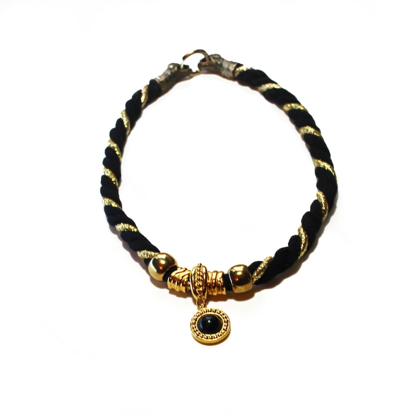 May include: A black and gold rope necklace with a gold pendant featuring a black stone. The necklace has a twisted rope design with gold accents. The pendant is round and has a gold frame.