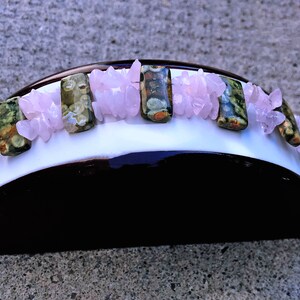 May include: A handcrafted headband featuring a white base with a dark brown underside. Adorning the headband are rectangular green and brown stones, interspersed with clusters of light pink crystals. The headband is designed for a comfortable fit.