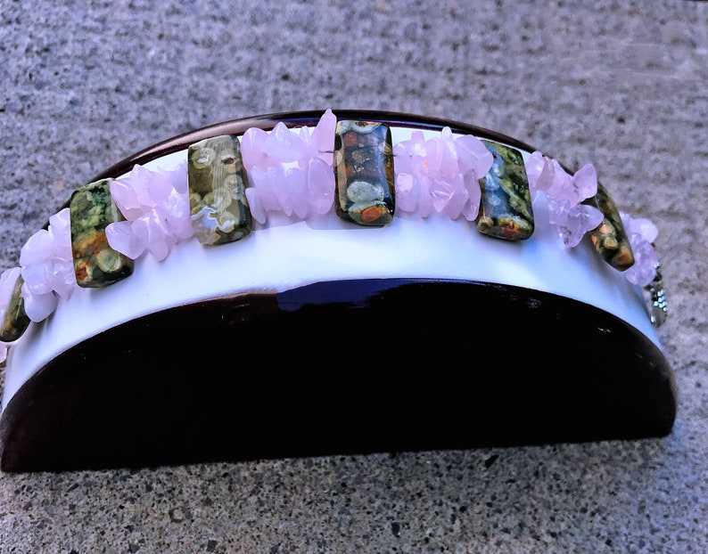 May include: A handcrafted headband featuring a white base with a dark brown underside. Adorning the headband are rectangular green and brown stones, interspersed with clusters of light pink crystals. The headband is designed for a comfortable fit.