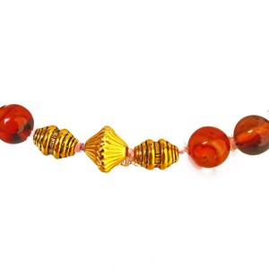 May include: A necklace featuring a strand of large, round, red and brown beads interspersed with gold-colored metal beads. The necklace is strung on a pink cord and is set against a white background.