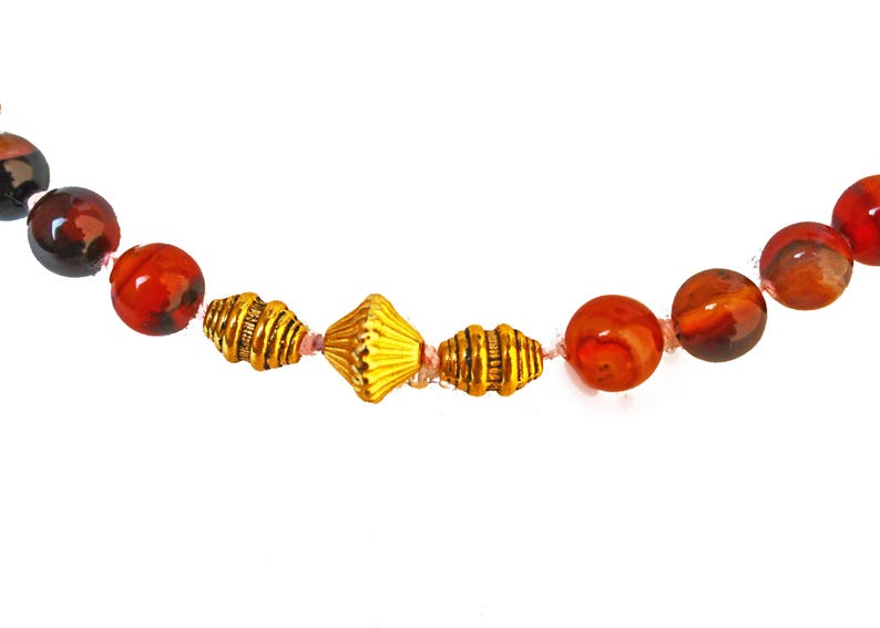 May include: A necklace featuring a strand of large, round, red and brown beads interspersed with gold-colored metal beads. The necklace is strung on a pink cord and is set against a white background.