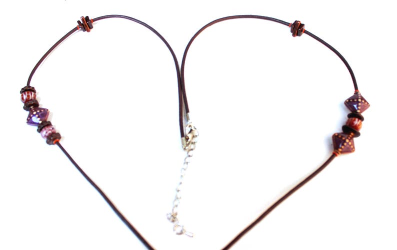 May include: A necklace with a dark brown cord and decorative beads. The beads include purple, brown, and orange elements. The necklace has a silver-colored clasp and a chain for adjusting the length.