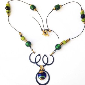 May include: A handcrafted necklace featuring a dark metal pendant with three interlocking circles and a blue and green glass bead. The necklace has green and gold beads strung on a dark wire, with a gold clasp.