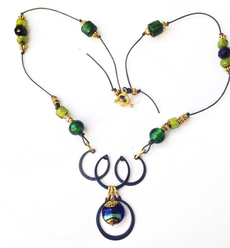 May include: A handcrafted necklace featuring a dark metal pendant with three interlocking circles and a blue and green glass bead. The necklace has green and gold beads strung on a dark wire, with a gold clasp.