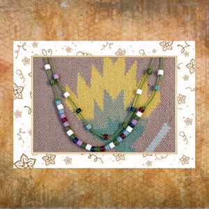 May include: A beaded necklace with a green cord features a variety of colorful beads, including white, blue, purple, and red. The necklace is displayed on a patterned background with a white border decorated with floral and swirl designs.
