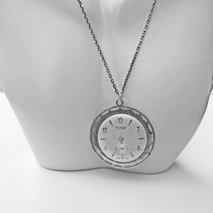 May include: A silver-toned pendant necklace featuring a vintage watch face. The watch face, marked "SUPER LUXE," displays the time with black numerals and hands. The pendant is suspended from a delicate chain, creating a classic and elegant accessory.
