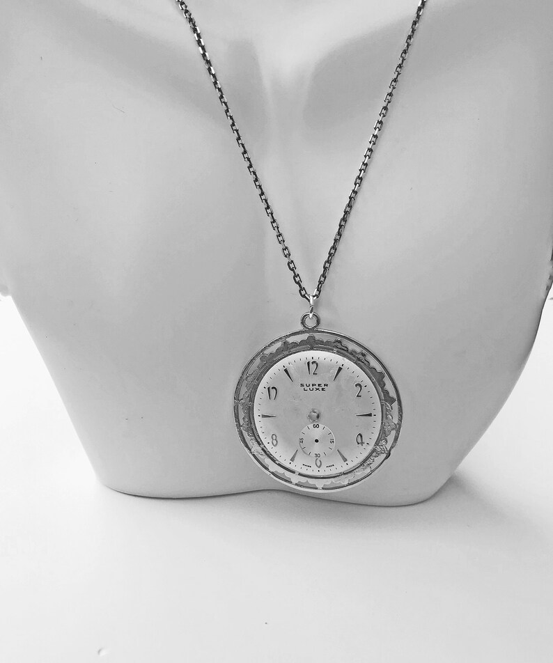 May include: A silver-toned pendant necklace featuring a vintage watch face. The watch face, marked "SUPER LUXE," displays the time with black numerals and hands. The pendant is suspended from a delicate chain, creating a classic and elegant accessory.