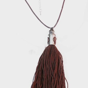 May include: A long necklace featuring a dark brown tassel pendant. The necklace has a brown cord and silver-toned metal accents. The upper portion of the necklace has decorative beads with red, white, and blue patterns.