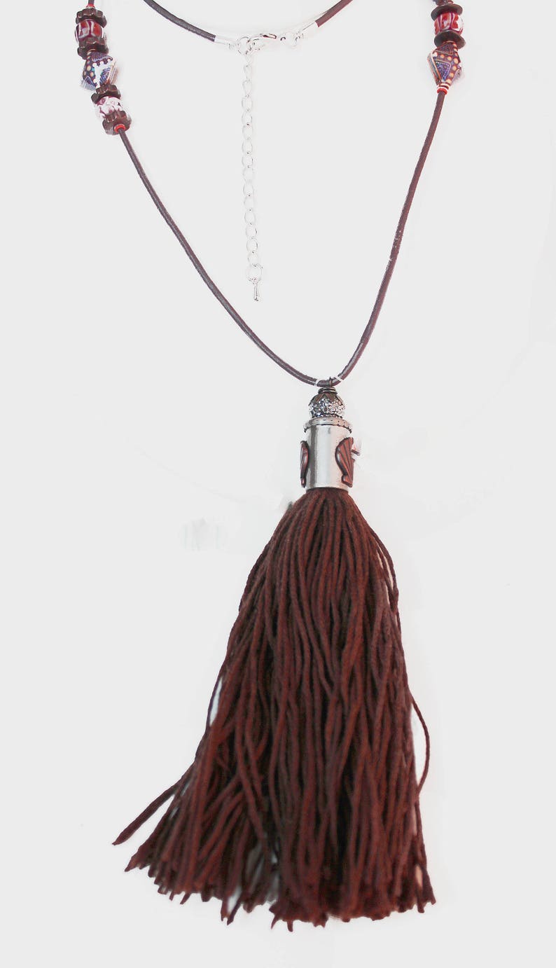 May include: A long necklace featuring a dark brown tassel pendant. The necklace has a brown cord and silver-toned metal accents. The upper portion of the necklace has decorative beads with red, white, and blue patterns.
