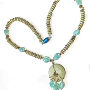 May include: A handcrafted necklace featuring a large, round, pale green pendant with a fan-like wire design and dangling aqua-colored teardrop beads. The necklace has a strand of small, light green beads interspersed with aqua-colored stones and brass accents. A blue oval clasp secures the necklace.