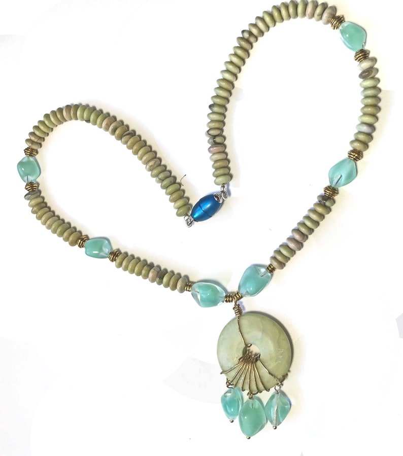 May include: A handcrafted necklace featuring a large, round, pale green pendant with a fan-like wire design and dangling aqua-colored teardrop beads. The necklace has a strand of small, light green beads interspersed with aqua-colored stones and brass accents. A blue oval clasp secures the necklace.