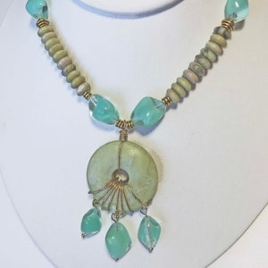 May include: A handcrafted necklace featuring a large, light green circular pendant with a central hole, accented by gold wire and dangling teardrop-shaped beads. The necklace includes light green and clear beads, and is displayed on a white bust.