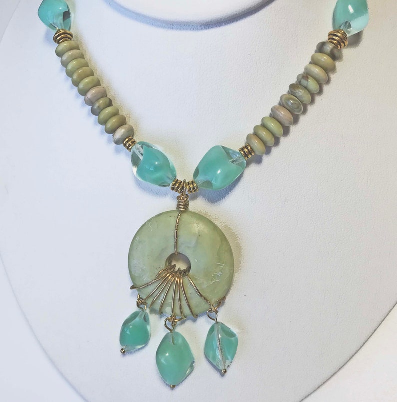 May include: A handcrafted necklace featuring a large, light green circular pendant with a central hole, accented by gold wire and dangling teardrop-shaped beads. The necklace includes light green and clear beads, and is displayed on a white bust.