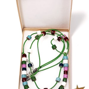 May include: A colorful necklace with a green cord and various colored beads, including white, red, blue, green, and purple, is displayed inside a beige box. The necklace has a silver clasp. A gold star is in the lower right corner.