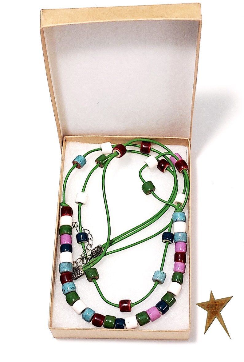 May include: A colorful necklace with a green cord and various colored beads, including white, red, blue, green, and purple, is displayed inside a beige box. The necklace has a silver clasp. A gold star is in the lower right corner.