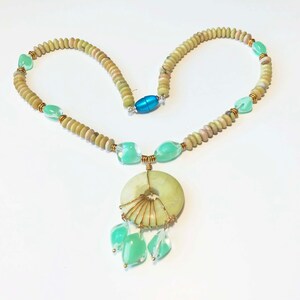 May include: A handmade necklace featuring a large, light green circular pendant with wire detailing and dangling green teardrop-shaped beads. The necklace has a strand of small, round, light brown beads, with accents of green and gold. A blue magnetic clasp secures the necklace.