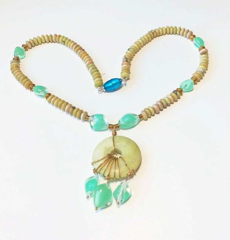 May include: A handmade necklace featuring a large, light green circular pendant with wire detailing and dangling green teardrop-shaped beads. The necklace has a strand of small, round, light brown beads, with accents of green and gold. A blue magnetic clasp secures the necklace.