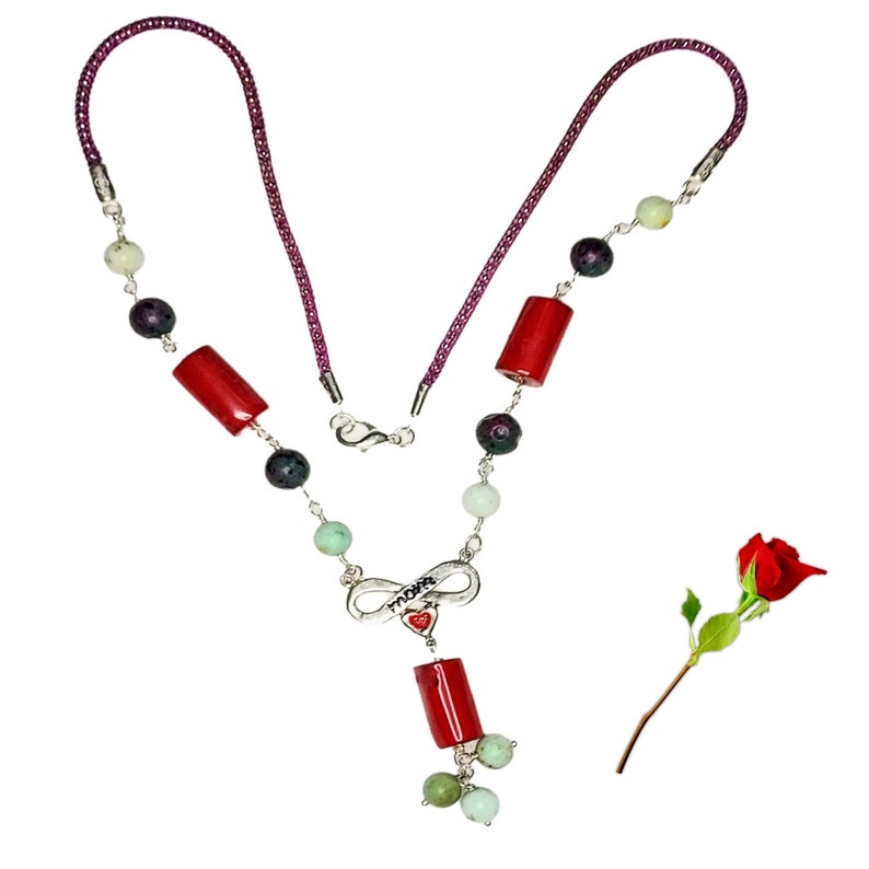 May include: A silver necklace with red coral beads, green beads, and a silver infinity charm with the word 'Mom' and a red heart. The necklace is on a white background with a red rose.