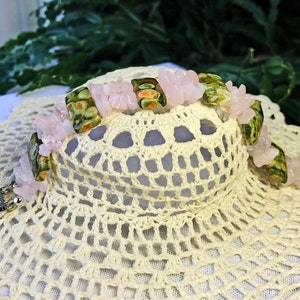 May include: A beaded bracelet featuring pink rose quartz chips and rectangular green and orange patterned beads. The bracelet has a silver clasp and is displayed on a cream-colored, crocheted surface.