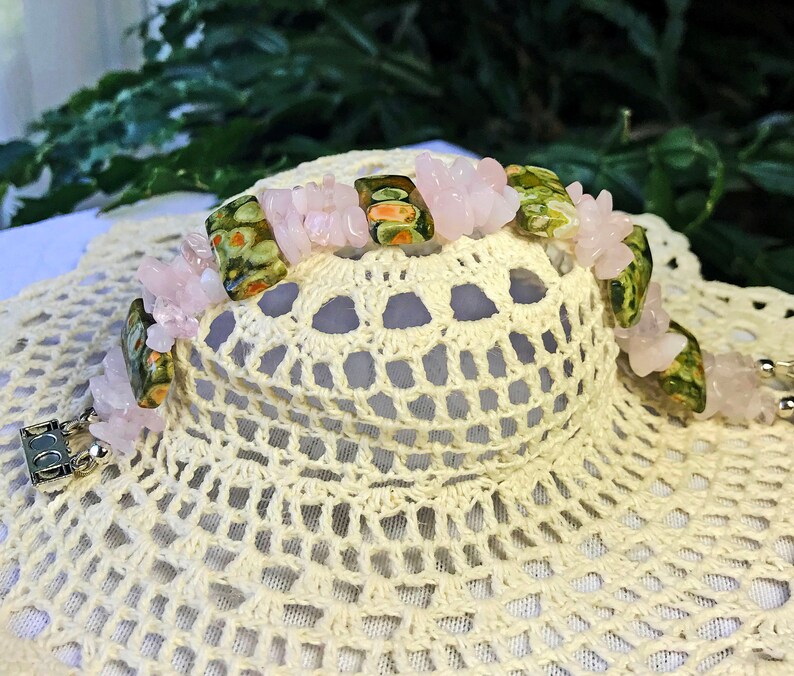 May include: A beaded bracelet featuring pink rose quartz chips and rectangular green and orange patterned beads. The bracelet has a silver clasp and is displayed on a cream-colored, crocheted surface.