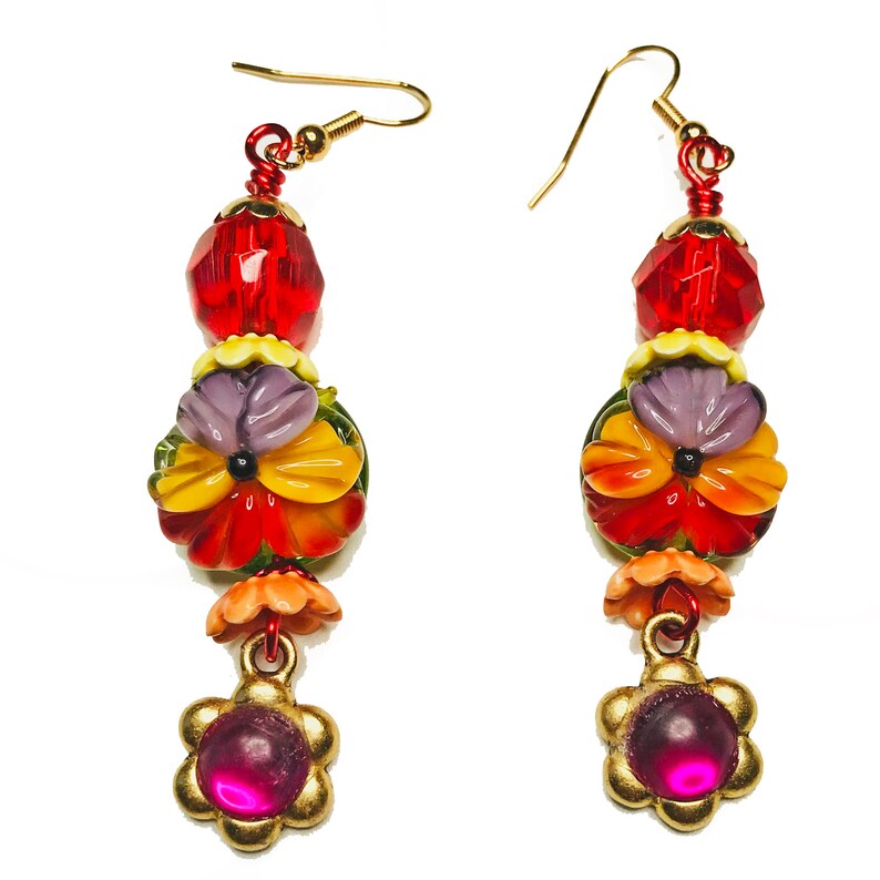 May include: A pair of dangling earrings with a floral theme. Each earring features a red faceted bead, a cluster of colorful flower beads in shades of red, yellow, and purple, and a gold-colored flower charm with a pink center. The earrings have gold-colored hooks.