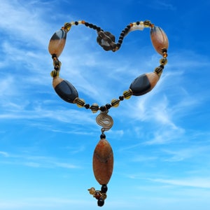 May include: A handcrafted necklace with a heart-shaped design, featuring large, oval-shaped beads in shades of brown and black, interspersed with smaller yellow and black beads. A spiral pendant and a long, oval-shaped pendant hang from the center. The background is a bright blue sky.