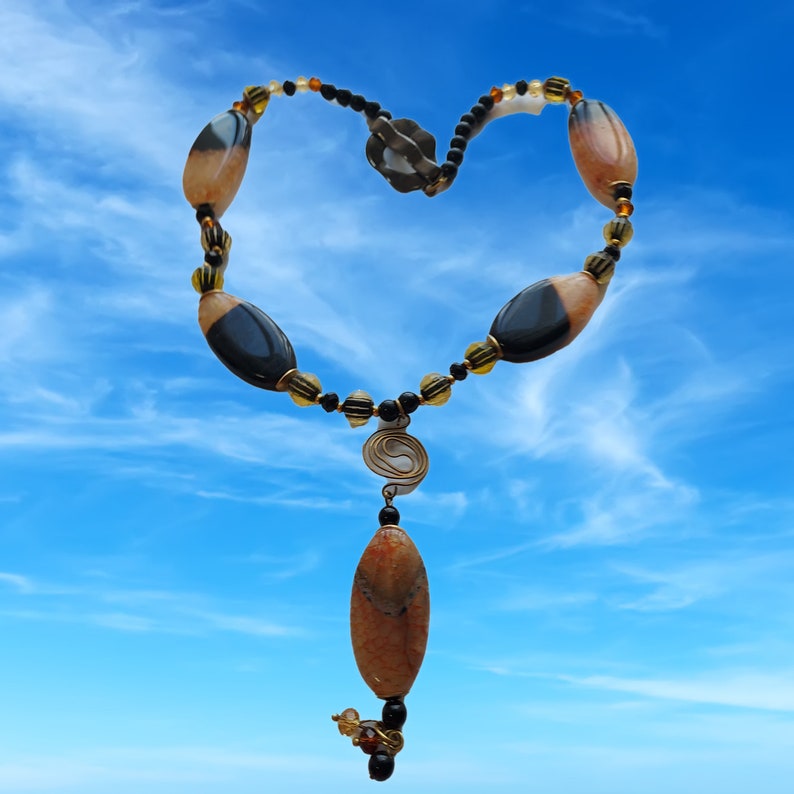 May include: A handcrafted necklace with a heart-shaped design, featuring large, oval-shaped beads in shades of brown and black, interspersed with smaller yellow and black beads. A spiral pendant and a long, oval-shaped pendant hang from the center. The background is a bright blue sky.