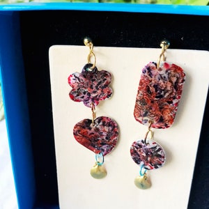 May include: A pair of unique earrings with a floral, heart, and rectangular design. The earrings feature a mix of red, purple, and white patterns. Each earring has a small gold charm at the bottom, and they are displayed on a white card.