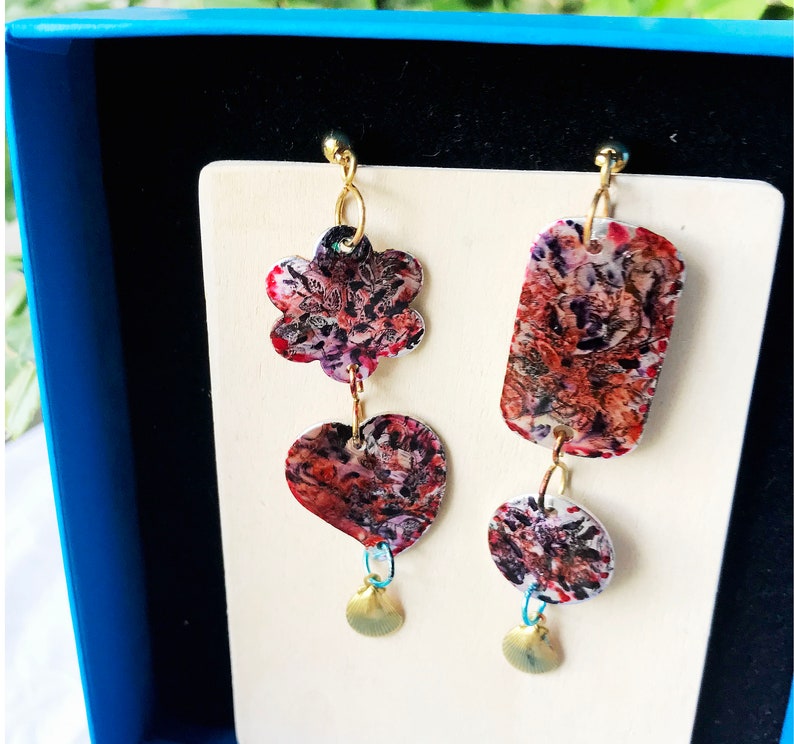 May include: A pair of unique earrings with a floral, heart, and rectangular design. The earrings feature a mix of red, purple, and white patterns. Each earring has a small gold charm at the bottom, and they are displayed on a white card.