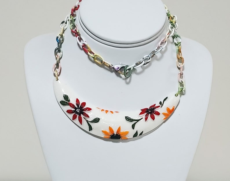 May include: A white, crescent-shaped necklace with hand-painted red and orange flowers and green leaves. The necklace is attached to a colorful chain with a lobster clasp. The jewelry is displayed on a white bust.