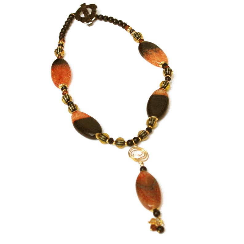 May include: A handcrafted necklace featuring oval-shaped, reddish-brown and black beads, interspersed with gold-toned accents. A gold-colored spiral pendant hangs from the center, with a matching oval bead at the bottom. The necklace is secured with a bronze-colored clasp.